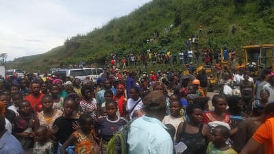 The image shows a large crowd of people gathered on a dirt road in Sake, Democratic Republic of Congo. The road is lined with parked cars and trucks, and a yellow construction vehicle is parked in the center of the image. The people are standing in a dense crowd, with many of them wearing casual clothing. The background shows a steep hillside covered with trees and vegetation. The crowd appears to be waiting or watching something, but there are no obvious signs of any activity. There is a dense group of people standing closely together in the foreground, with more people on the hillside in the background. The crowd is mostly composed of women and children, with a few men scattered throughout. There is a man in the center of the image, wearing a blue shirt and a backpack, standing in front of the crowd. The image is taken from a low angle, giving the viewer a perspective of looking up at the crowd. It appears to have been taken on a sunny day, with a clear blue sky above. The scene is seemingly taking place in a rural part of Sake, given the dirt road and the surrounding vegetation. The overall scene is one of anticipation and uncertainty, as the crowd appears to be waiting for something or observing an event.