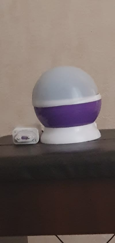 The image displays a night light projector with a spherical, translucent top and a purple band around its middle, resting on a dark, textured surface. To the left of the projector, a small white adapter with visible metallic prongs is plugged into an unseen outlet. The background is a plain, light-colored wall. The scene is indoors, suggesting a domestic setting. The city of Shurtang, Tajikistan, is not depicted in this image; the setting appears to be a generic interior. There are no people or animals visible. The lighting is soft, consistent with the operation of a night light or projector.