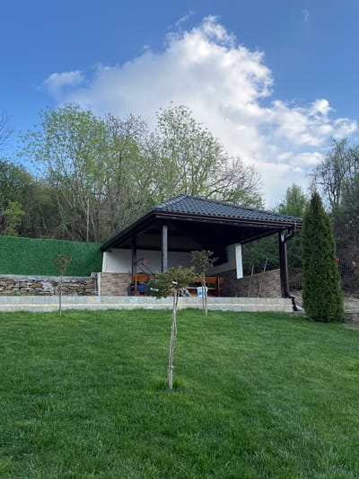 The image shows a well-maintained backyard in Kribul, Bulgaria. A structure, possibly a summer kitchen or gazebo, with a dark roof and light-colored walls stands in the center. There's a stone or brick base around the perimeter of the structure and the property, bordering a lush green lawn. A tall, thin evergreen tree is present near the corner of the structure. Deciduous trees are visible in the background. The sky is blue with scattered white clouds. No people are visible in the frame.