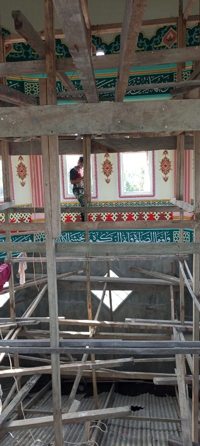 This vertical image depicts the interior of a building undergoing construction or renovation in Kumbang, Indonesia, identified by its Islamic decorative motifs and Arabic calligraphy, suggesting it is a mosque. The scene is largely filled by an extensive, multi-level scaffolding system composed of unpainted, rough-hewn wooden beams and planks, occupying the lower two-thirds of the frame and extending upwards.

In the upper third of the image, a decorated interior wall section is visible. It features three rectangular windows with white frames, through which bright natural light enters and green foliage is discernible outside. The light cream-colored wall surfaces between and beside these windows are adorned with symmetrical, ornate patterns in red, gold, and green. Below the windows, a horizontal band displays geometric motifs in red, gold, and green. Further down, a prominent teal-colored band features white Arabic calligraphy, beneath which is a smaller band with a green and white geometric pattern. The upper wall or ceiling area above the windows is painted green with intricate light-green and yellow patterns, from which a single white light bulb is suspended.

One individual is observed in the upper section, standing on a higher beam or platform between the left and central windows. Dressed in a patterned short-sleeved shirt and dark pants, the person faces right and appears to be engaged in work on the decorated wall, with their right hand positioned near its surface.

On the mid-left side of the wooden scaffolding, several items of clothing (one red, others light-colored) hang from a horizontal beam, accompanied by a length of rope. Below the dense wooden framework, a corrugated metal roof or floor is visible, showing signs of wear and several open sections that expose dark spaces beneath. The environment is brightly illuminated by natural light entering through the windows.