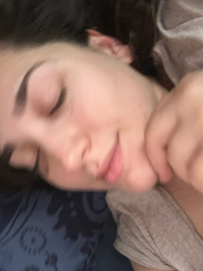 The image captures a close-up of an individual, identified as likely an adult female, resting in an indoor setting within Ancenis, France. Her eyes are closed, and a subtle smile is present on her lips, suggesting a state of sleep or deep relaxation. The individual possesses fair skin, with visible dark eyebrows and dark hair partially obscuring the top of the frame. A hand, presumably her own, supports or rests near her jawline and upper neck on the right side. She is dressed in a light grey short-sleeved garment, possibly a t-shirt. Her head is supported by a dark blue fabric object, likely a pillow, adorned with a lighter, indistinct patterned design. The scene is illuminated by soft, ambient lighting.