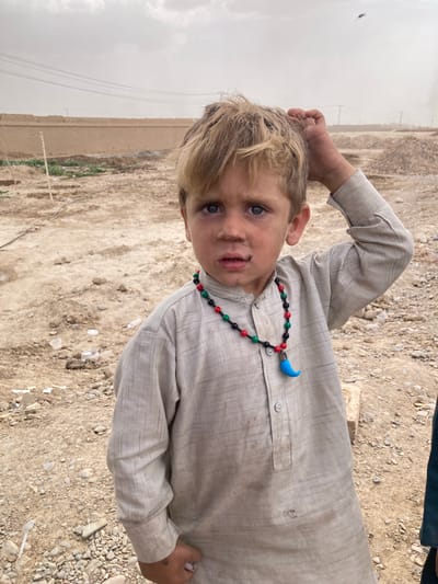 A young, blonde-haired boy with blue eyes stands on a dirt and gravel surface. He wears a light-colored shirt with a colorful beaded necklace featuring a crescent-shaped pendant. The boy has dirt on his face. In the background, a wall is visible, and a distant landscape fades into a cloudy sky. The location is in the city of Saranan, Pakistan.