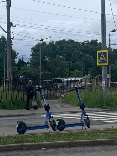 The image shows an outdoor scene in Рассудово, Russia. Two electric scooters, one blue and one dark, are parked on the side of a road next to a crosswalk. The blue scooter has the text "MXG578" on it, and the other scooter has "MX184E". In the background, there is a pedestrian crossing sign, a metal fence, and lush green trees. A person in dark clothing is partially visible, appearing to be walking away from the scooters. Overhead power lines are prominent in the sky, which is overcast, suggesting a cloudy day. The road surface is visible, as is a strip of grass with small white flowers bordering the road. The scene appears to be a public area, possibly a park or a street with pedestrian amenities.