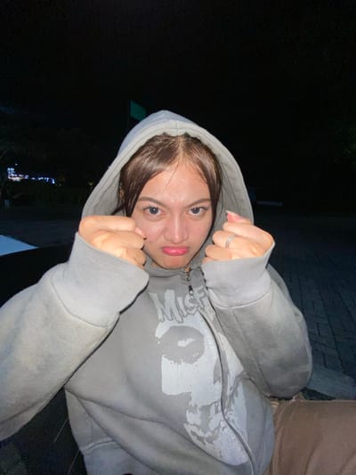 A young woman is captured in a close-up, nighttime portrait outdoors, with a dark background suggesting a city setting. She is wearing a grey hooded sweatshirt, zipped up, with a white graphic design on the front. Her hair is brown and tied back, with strands framing her face. She is making a pouting expression and has her fists clenched near her face, appearing to be in a playful or defiant pose. The lighting appears to be artificial, casting a slight glow on her face and clothing. The surroundings suggest a paved area, possibly a street or walkway, with some faint lights visible in the distance on the left side of the frame. There is no explicit indication of the location being in Mekar Bakti, Indonesia.