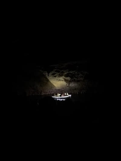 The image shows a dimly lit cave in Manacor, Spain. A guided tour boat is visible with several people onboard, illuminated by small lights. A beam of light shines on a rock wall next to the boat. Stalactites are seen on the cave ceiling. The surrounding environment is dark and rocky.