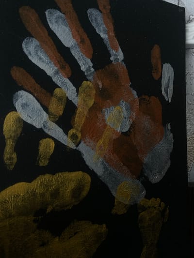 The image shows a close-up view of handprints and footprints made with paint on a black surface, possibly a canvas or poster board. The prints are in metallic gold, silver, and a rusty brown color. Some prints overlap. The texture of the paint appears somewhat thick and textured, revealing brush strokes or the texture of the surface it was applied to. The lighting suggests the artwork is in a dimly lit indoor environment. There is no indication of a specific location within the city of Пешнево, Russia, as the image focuses solely on the artwork.
