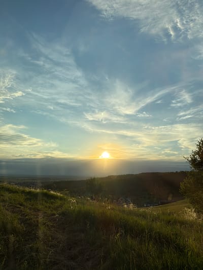 This is a vertical, landscape-oriented photograph taken in Raìmanovo, Russia. The image depicts a serene sunset scene with a bright sun low on the horizon, casting a warm glow across the sky. Wispy white clouds are scattered across a pale blue sky, creating a soft, ethereal atmosphere.
In the foreground, lush green grass, partially illuminated by the setting sun, lines the right side of the frame. The grass appears tall and somewhat unkempt, with some lighter-colored seed heads visible. Beyond the grassy field, a dark, forested hillside slopes down towards the horizon. Scattered houses with white roofs can be seen nestled in the valley below the hillside, suggesting a rural or semi-rural setting.
The overall mood of the image is peaceful and tranquil, evoked by the soft light, the natural landscape, and the quiet beauty of the sunset. The time of day is clearly sunset, indicated by the sun's position and the warm, diffused light. The weather appears to be clear with some high-altitude clouds. There is no visible text in the image.
