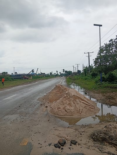 The scene depicts a road in Igrita, Nigeria, under an overcast sky. A large pile of tan-colored sand sits on the side of the road, partially submerged in a puddle of water. More puddles and mud are visible on the road surface and surrounding ground. To the left, a man in an orange shirt walks along the edge of the road. In the background, there is a flatbed truck, cranes, and various buildings under construction. Power lines and utility poles run along the right side of the road, near some vegetation. A modern street light stands to the right.
