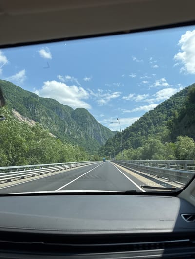 The image shows a view from inside a car driving on a highway through a mountainous, forested region. The highway is paved with asphalt and has white lane markings. It is bordered by a metal guardrail on both sides. Lush green trees cover the steep slopes of the mountains that flank the highway. The sky above is blue with scattered white clouds. A few streetlights are visible along the side of the road. The dashboard of the car is visible in the foreground. There are no people visible in the image, and no specific landmarks are identifiable that would suggest the location within the city of Monastyr, Russia, as the image depicts a natural landscape outside of a city.