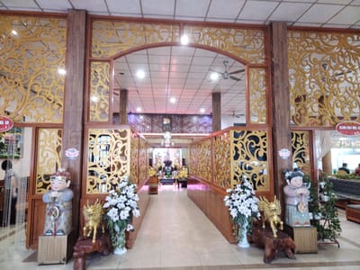 The image depicts a view of the interior of a building, possibly a temple or religious structure, located in Chu Văn An Ward, Vietnam. The scene is framed by an elaborate wooden entryway with ornate gold-colored filigree designs. Two child-like statues stand on either side of the opening, along with flower arrangements, a sculpture of a golden kirin and various potted plants. The statue on the left is a male figure and the statue on the right is a female figure.

The interior beyond the entryway features a long corridor with similar wooden dividers and floral decorations. In the distance, religious icons and objects are visible. The ceiling is lined with fluorescent lights. The building appears to be well-maintained and illuminated, suggesting active use and care. The setting includes tiled floors, and the space is open and seemingly spacious.

A few people are visible in the far background, near what appears to be the entry to the building behind the ornate decorations.