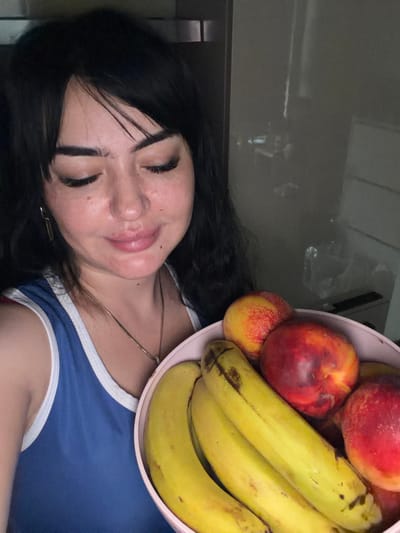 A young woman with dark hair and bangs is holding a pink bowl filled with fruit. She is wearing a blue tank top with white trim. Her eyes are closed, and she appears to be smiling. The bowl contains bananas and peaches. The background is out of focus, but appears to be an indoor setting, possibly a kitchen. There are reflections in a glassy surface behind her, suggesting she may be near a refrigerator or a window. No specific landmarks or details place this scene in İzmit, Turkey.
