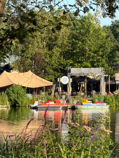 In Heijen, Netherlands, a calm body of water reflects the surrounding greenery and a bright sky. On the water, two miniature boats are docked. The boat closer to the viewer is predominantly red, with white accents and lettering reading "KIDS HAVEN LINES SERVICE." The second boat, to its right, is light blue with red and yellow detailing, featuring the name "The Blue Fisher" on its side. Behind the boats, a beige tent provides shade over an outdoor seating area. Beyond the tent and seating area, lush green trees form a dense backdrop. In the foreground, purple wildflowers with green stems grow along the water's edge, adding a touch of color to the scene. The overall impression is one of a serene, outdoor recreational area.