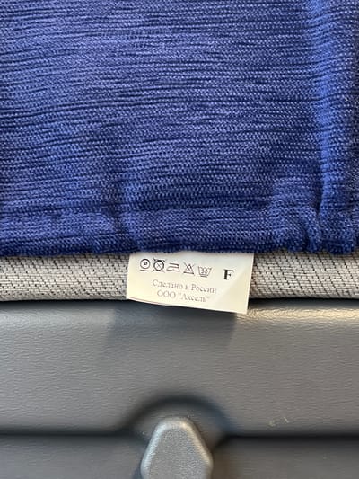 The image shows a close-up of a dark blue textured fabric, possibly clothing, with a white care label attached to it. The label features several laundry care symbols: a "P" in a circle (indicating dry cleaning is permitted), a crossed-out triangle (meaning do not bleach), a crossed-out basin with a hand (indicating do not wash by hand), a basin with "30" inside (indicating machine wash at 30 degrees Celsius), and the letter "F" (indicating dry cleaning with any solvent except trichloroethylene). Below the symbols, text in Russian reads "Сделано в России" which translates to "Made in Russia" and "ООО 'Аксель'" which is likely the company name "OOO 'Aksell'". The fabric is draped over a textured grey surface, which is then placed on a dark grey, slightly reflective surface that appears to be a plastic or leather material, possibly the interior of a vehicle or furniture. The background is out of focus and dark. There are no people visible in the image. The scene does not provide any specific information that would allow for localization within the city of Lomonosov, Russia.