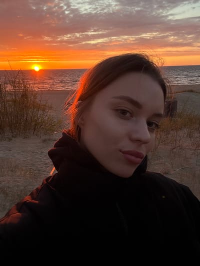 A portrait of a woman is set against the backdrop of a sunset over the sea. The woman, positioned on the right side of the frame, has light skin, brown hair, and is wearing a dark jacket. The background reveals a seascape with the sun near the horizon, creating an orange-yellow glow. The scene appears to be a beach setting with some sparse vegetation. The location is Ādaži, Latvia, based on geographical recognition.
