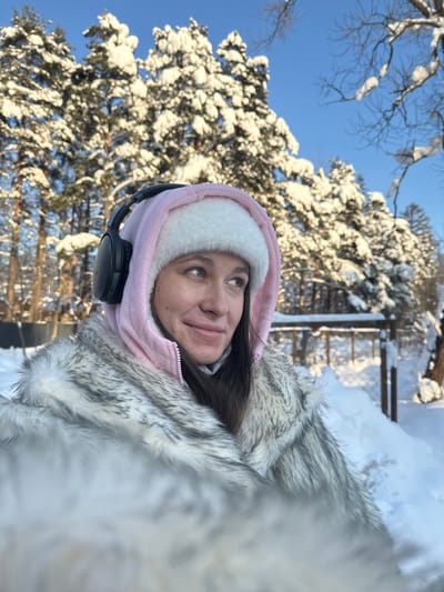 A young adult female is depicted outdoors in a snowy environment, identified as Дубки, Russia. She is shown from the upper chest upward, looking towards the upper right of the frame with a slight smile. She wears a white, furry winter hat under a pink hooded fleece jacket, with black over-ear headphones placed on top of the hood. A grey and white faux fur coat is visible on her shoulders. Her dark hair extends beneath the hat. The background features a deeply snow-covered landscape under a clear blue sky. Numerous coniferous trees, heavily laden with snow, dominate the mid-ground and background. Bare deciduous tree branches, also dusted with snow, are visible on the right. A wooden fence, partially obscured by snowdrifts, is present in the middle ground to the right. The ground is uniformly covered in deep, white snow, with shadows suggesting bright sunlight.