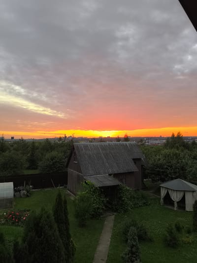 This image depicts a rural or suburban landscape in Russia at sunset. The sky is a prominent feature, transitioning from soft greys at the top to vibrant oranges and reds along the horizon, indicating the sun is setting. Wispy clouds are visible throughout the sky.

The foreground is dominated by lush green grass and various trees, including tall conifers and dense bushes. Several man-made structures are present: a weathered wooden house with a dark, sloped roof sits centrally, with a smaller attached covered porch. To the left, a small greenhouse is partially visible. To the right, a light-colored gazebo with draped curtains offers a sheltered seating area. A dark wooden fence runs along the back of the property.

In the mid-ground, more trees form a thick line, beyond which a distant town or industrial area can be seen with buildings and structures. Power lines and utility poles are also visible in the distance.

The overall scene evokes a sense of tranquility and the end of a day. The vibrant sunset colors suggest warmth and beauty, contrasting slightly with the shadowed foreground and rustic structures. There is no visible text in the image.