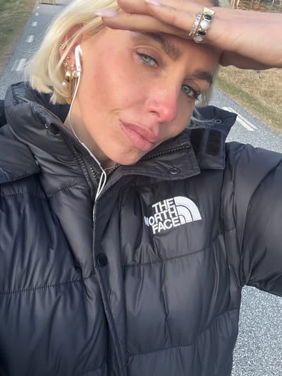 A woman is facing the camera while standing in Strömstad, Sweden. She has blond hair and is wearing a black North Face puffer jacket. The woman has multiple rings on her fingers and several earrings. She is also wearing white headphones. The background appears to be a paved road and vegetation.