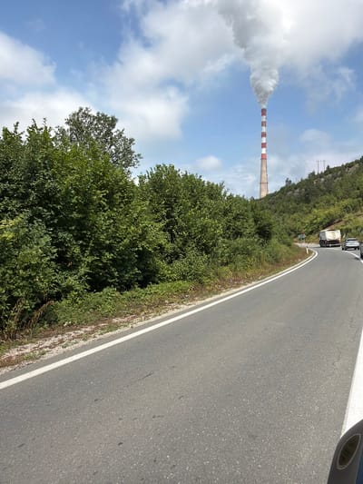 A daytime, medium-shot, high-angle photograph captures a rural road curving through lush green foliage in Zabrđe, Montenegro. A tall, red and white striped smokestack dominates the background, emitting a thick plume of white smoke that disperses into a blue sky dotted with white clouds. The road, paved with asphalt, is marked with a white line and shows signs of wear. A white truck and a silver car are visible on the road, traveling away from the camera. The surrounding environment is characterized by dense vegetation, including trees and bushes, with a hillside visible in the distance. The scene appears to be a snapshot of a typical day in the Zabrđe region, showcasing the interplay between industrial activity, natural landscape, and human infrastructure.