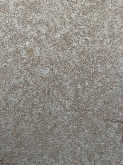 The image shows a close-up of a textured surface, likely wallpaper or a similar wall covering. The texture is irregular, with raised ridges and valleys, creating a tactile appearance. The color is predominantly a light beige or taupe, with some areas appearing lighter due to the texture and lighting. Scattered throughout the surface are small flecks of what appear to be glitter or metallic particles, adding a subtle shimmer. The background is a uniform, muted tone. There are no people, objects, actions, or interactions visible in this image. The context provided regarding Beryozovka, Russia, is not visually represented in the image itself, as it solely depicts a textured surface.