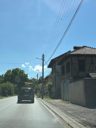 The image shows a road with a car driving away from the camera in the city of Beli Osam, Bulgaria. The sky is blue with a few white clouds. Overhead, power lines stretch across the scene, with a small bird perched on one of them. To the right of the road, there is a building with a tiled roof and a distinctive timber-framed facade. Further down the road, more houses and trees are visible. The car, a dark-colored hatchback, has a license plate that reads "PA 3063 BT". The road surface is asphalt, and there is a concrete curb and a gravel shoulder along the side. The overall scene depicts a rural or suburban street in daylight.