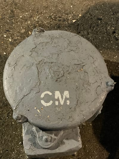 The image shows a close-up, high-angle shot of a round, gray, metal cover set into the ground. The cover is weathered, with peeling paint and visible cracks. On the surface of the cover, the white letters "CM" are stenciled. A circular emblem is also present but is indistinct due to the condition of the paint. The cover is mounted on a rectangular concrete base, which also appears weathered. The surrounding ground is rough and appears to be a mix of dirt, small stones, and some debris. The lighting suggests it may be nighttime or a dimly lit area. There are no people or animals visible in the image. The location is specified as Samur, Russia.
