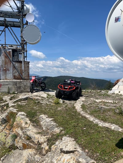 The image depicts a scenic overlook with two off-road vehicles parked on a rocky, grassy plateau. A red and white dirt bike is parked to the left, closer to a weathered, industrial-looking structure with large satellite dishes mounted on a metal tower. To the right of the dirt bike, a black and orange all-terrain vehicle (ATV) is parked on a winding stone path. The background features a vast, forested mountain range under a bright blue sky with scattered white clouds. The location is identified as Simeonovets, Bulgaria, suggesting it's a natural or recreational area. The scene is static, with no people or visible activity other than the parked vehicles.