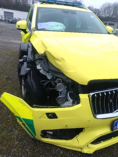 A severely damaged yellow emergency vehicle is depicted in Bitburg, Germany, following a front-end collision. The vehicle, appearing to be a Volvo XC90 SUV, likely functions as an ambulance or rescue unit based on its yellow livery and rooftop blue emergency light bar. Extensive damage is concentrated on the front left side. The hood is severely crumpled and buckled. The front bumper is detached on the left and rests on the ground, revealing internal mechanical components and a distinct green stripe on its separated left segment. The left front fender is missing, exposing the vehicle's internal structure and horn assembly. The driver's side airbag is deployed and visible on the dashboard, alongside several white papers. The right side of the front grille appears largely intact, with a partial license plate starting with 'B' visible beneath it. The vehicle is stationary on a dark gravel surface. In the background, industrial-style buildings, a fence, and bare trees are discernible under an overcast sky, indicating an outdoor lot or incident scene. No people are visible.