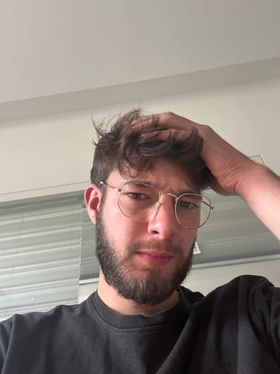 An adult male with light skin, dark wavy hair, a beard, and wire-rimmed glasses is depicted from the chest up, positioned centrally in the frame. His left hand is raised, with fingers touching the top of his head, ruffling his hair. He wears a dark, possibly black, t-shirt. The man looks directly into the camera with a neutral expression. The background consists of a plain, light-colored ceiling or upper wall area occupying the top portion of the image. To the left and right, behind the subject, visible elements include horizontal light-colored window blinds or slats. The scene is illuminated by ambient light. This image is documented in Lièpvre, France.