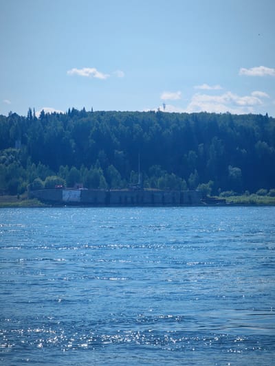 The image shows a wide, calm body of water, likely a river, with a textured surface reflecting the sunlight. In the background, a dense, dark green forest covers a hilly landscape under a clear blue sky with scattered white clouds. Along the shoreline, a long, low concrete structure, possibly a pier or a wall, is visible. This structure has several arched openings or recessed areas and appears aged, with some discoloration and repairs. On top of this structure, there are a few indistinct objects, including what might be machinery or buildings, and some scattered vegetation. A white pole or antenna is also visible extending upwards from the structure. The overall scene depicts a serene, natural environment with a man-made element integrated into the landscape. This is likely the riverfront area of Абалаково, Russia.