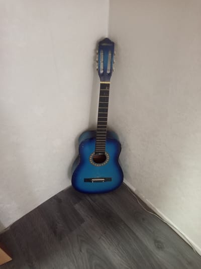 A blue acoustic guitar is leaning in the corner of a room against a white wall. The guitar has a dark fretboard with silver tuning pegs. The body of the guitar is a bright blue with a wooden soundhole. The floor is made of dark wood laminate. The image does not provide any information about the city of Hlusk, Belarus.
