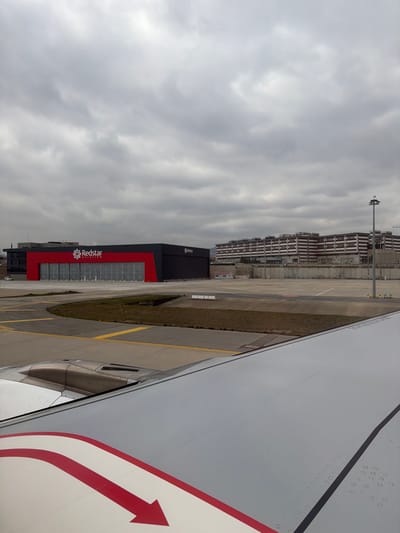 The image displays an exterior view from the right wing of an aircraft, positioned on an airfield apron at Sabiha Gökçen International Airport, located near Tuzla, Turkey. The aircraft's white wing, featuring a red arrow decal pointing aft-outboard and a section of its engine nacelle, occupies the lower right quadrant. Directly ahead, a large, modern hangar building belonging to "Redstar Aviation" is visible, characterized by a black structure with a prominent central red facade displaying the company's white logo and name. This building has multiple large, closed hangar doors. The foreground and midground consist of a grey asphalt taxiway or apron, marked with yellow lines, and interspersed with patches of dry, brown grass. In the background on the right, a multi-story industrial or commercial building complex, light brown in color with numerous horizontal bands and windows, is present. A tall, grey light pole stands in front of this complex. The sky is uniformly overcast with dense grey clouds. No individuals are visible, and the scene appears static.