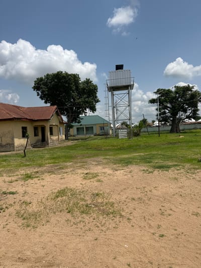 This media file depicts an outdoor scene under a bright, partly cloudy sky, likely in Lau, Nigeria. The image is oriented vertically. In the foreground, a patch of dry, sandy ground with sparse green grass occupies the right side. To the left of the sandy area, a grassy field stretches towards some buildings and a prominent water tower.

The central subject is a tall, metal water tower with a rectangular tank at the top, supported by a lattice structure. Adjacent to the water tower, a two-story building with a light green roof and yellow walls is visible. In front of this building, a smaller, older building with a rusty, corrugated metal roof and yellow walls is also present. A large, mature tree with dense dark green foliage stands between these two buildings, casting a shadow.

In the sky, several fluffy white clouds are scattered across a clear blue expanse. A small bird is visible in flight in the upper left portion of the image. The lighting suggests it is daytime, with the sun casting distinct shadows. The overall impression is of a functional, possibly institutional, setting with elements of nature. There is no visible text in the image.