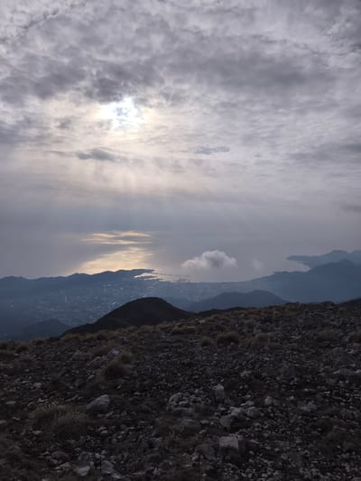 The image depicts a vast landscape observed from a high vantage point in Mali Mikulići, Montenegro. The immediate foreground consists of a rocky, uneven terrain covered with dark grey stones and sparse patches of dried, yellowish-brown grass. Beyond this rocky expanse, a series of darker mountain ridges descend towards a coastal plain. A densely populated urban area, characterized by numerous buildings, occupies the mid-ground, bordering a large body of water. A prominent port or harbor with visible piers extends into this body of water.
The water's surface displays a distinct, elongated reflection of bright light, originating from a partially obscured sun positioned in the upper-left quadrant of the overcast sky. The sky itself is covered by a thick layer of grey clouds, with visible rays of light piercing through the cloud cover and illuminating sections of the sky and water. A single, isolated white cloud floats above the water to the right of the primary sun reflection. In the distant background to the right, additional mountainous landforms frame another inlet or bay. No human figures or discernible actions are present within the scene.