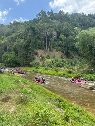 The image shows a river scene in Tambon Song Phraek, Thailand, filled with multiple rafts. A vibrant green forest covers the land on the left. The water is murky, and the rafts are spaced out along the river's length. Some rafts are pink, others orange or black, each carrying several individuals. People appear to be paddling. The landscape is lush with vegetation, and the sky shows a patch of blue.