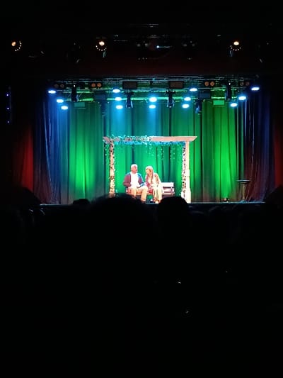 The image shows a stage with a man and a woman standing in the center. The man is wearing a brown shirt and the woman is wearing a white dress. They are positioned in front of a wooden structure adorned with greenery. The stage is lit with bright lights and the background is a dark green curtain. The image was captured in a theatre in General Pacheco, Argentina at coordinates -34.457, -58.633. There is a crowd in the foreground, but the camera is focused on the stage, and their faces cannot be seen. The audience is obscured by darkness. The scene is a performance taking place in a theatre.