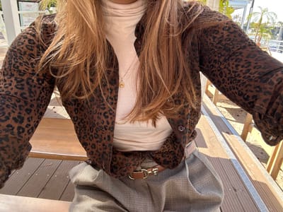The image presents a close-up, chest-down view of an individual seated outdoors in Levallois-Perret, France. The person wears an unbuttoned brown and black leopard-print shirt over a light beige turtleneck top. Grey pleated trousers are visible below the waist, secured by a thin brown leather belt with a gold-colored buckle featuring a horse-head design. A delicate gold chain necklace with a small circular gold pendant is visible on the turtleneck. Long, reddish-brown hair cascades over the individual's shoulders and chest. The subject is seated on a light brown wooden slatted bench, with a similar wooden deck interspersed with metallic strips visible on the ground. The background, under clear daylight, shows green foliage, including elements resembling palm trees, and parts of modern structures, one with a white sign displaying red letters, possibly "HLN."