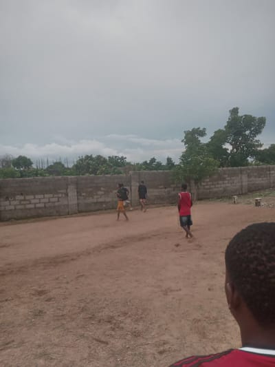 The image captures a dirt field in Achara Ndiagu, Nigeria. A low wall runs along the left side of the field. Four boys are present, seemingly barefoot. One boy wears a red shirt and dark shorts. Two others are further away, one in a black shirt and shorts, and another in a black shirt and yellow shorts. The fourth boy is partially cropped in the foreground with only his hair and shoulder visible, wearing a red shirt. The sky is overcast, and there are trees behind the wall.