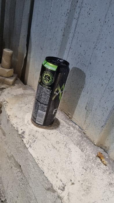 A black and green energy drink can is positioned horizontally, casting a shadow on a textured grey surface above. The can is slightly tilted, resting against a light grey concrete structure. A bolt with nuts is visible near the bottom left of the frame, and a small, dried leaf sits near the top right. The background is composed of various shades of grey and white concrete textures.