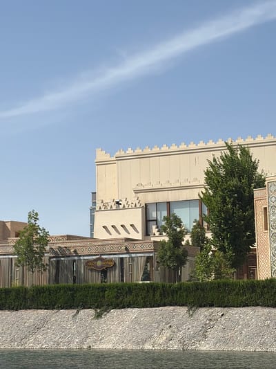 The image captures a vibrant daytime scene in Kuchtumgali, Uzbekistan, showcasing architectural details, lush greenery, and water. The perspective is from a slightly elevated position, looking across a water feature towards buildings.
In the foreground, a textured, light gray wall or embankment, composed of small pebbles, runs horizontally, bordering a calm body of water to its right. Beyond this, a neatly trimmed green hedge provides a natural boundary.
The central focus is on two prominent buildings. The taller, lighter-colored structure on the left features a distinctive stepped, castellated roofline, reminiscent of traditional fortresses or palaces. Its walls are smooth, and it has several large, rectangular windows. A lush green tree with dense foliage partially obscures this building.
To the right and below, another building displays intricate patterns on its facade, characteristic of traditional Uzbek tilework or brickwork designs. Attached to this building, a dark, ornate sign with gold or cream lettering clearly reads "OLOT SOMSA," indicating a restaurant or cafe specializing in local cuisine. More trees with green leaves are visible around this lower building.
The upper left portion of the image is dominated by a clear, light blue sky, featuring long, thin, wispy white clouds or contrails stretching diagonally across it. The bright, even lighting suggests a sunny day, likely late morning or early afternoon. No people are visible in the scene, which appears peaceful and well-maintained.