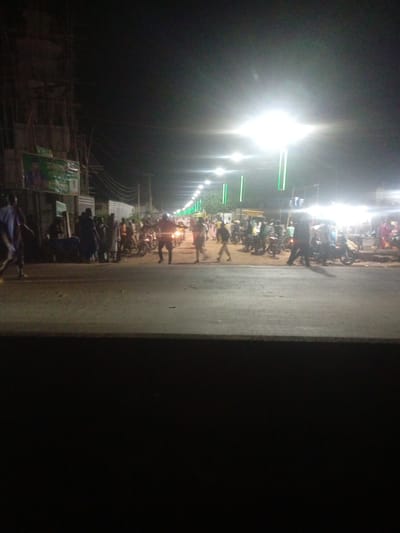 This media file captures a nighttime scene on a street in Tundun Wada South, Nigeria. The image is illuminated by bright streetlights, some of which are accented with green vertical lights. The street is paved and appears to be busy with people and motorcycles.
The subjects include a crowd of individuals, some of whom are gathered in groups while others are walking or standing. Several motorcycles are visible, suggesting they are a mode of transportation or perhaps part of a gathering. In the background, there are buildings and structures, one of which appears to be under construction with scaffolding. A banner or billboard with green coloring and some visible text is also present on a building to the left.
The activity appears to be a social gathering or a busy evening market/street scene. People are milling about, indicating a lively atmosphere. The time of day is clearly night due to the artificial lighting and the dark sky. No specific weather conditions are discernible, but the lighting suggests it is likely dry. The presence of multiple people and vehicles points to a populated urban or semi-urban area. There are no strong indicators of specific emotions, but the general scene suggests activity and potentially social interaction. The visible text on the banner is too blurry to be deciphered.