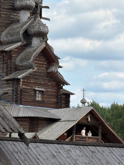 In Nevskii Parkleskhoz, Russia, a photograph captures a view of a historic wooden church against a cloudy sky. The church's architecture features multiple onion domes with shingled roofs, intricate wooden carvings along the eaves, and log construction. In the mid-ground, two individuals, a man in a white t-shirt and a woman in a light blue top, are visible on a wooden balcony, looking out from under the eaves of a building that appears to be part of the same complex. A cross is visible atop a smaller dome in the background, surrounded by lush green trees. The foreground is dominated by overlapping wooden roofs of varying pitches.