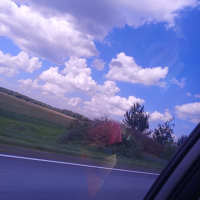 A wide-angle, slightly blurry shot captures a landscape viewed from a moving vehicle, likely a car, looking out the passenger side window. The scene is dominated by a bright blue sky filled with fluffy white and purple-tinged clouds. In the middle ground, a sloping field of green grass and brown earth stretches diagonally across the frame, bordered by a dense line of trees. The foreground consists of a dark asphalt road with a white line marking its edge. The right side of the frame shows the interior of the car, including a dark window frame and reflections on the glass. There are no people visible in the image. The overall impression is one of a rural landscape viewed during daylight, possibly late afternoon given the cloud colors.