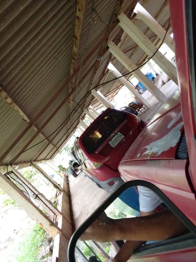 The image shows two red cars parked under a corrugated metal roof supported by wooden beams. The roof appears to be part of an open-air structure, possibly a carport or workshop. The first car, a hatchback, is visible on the left, and its license plate reads "AG987SA". The second car, a sedan, is on the right, and its hood is covered in patches of primer and rust. In the foreground, a portion of a side mirror reflects an arm and shoulder of a person wearing a white shirt. The surrounding environment is overgrown with lush green vegetation, suggesting a rural or semi-rural setting, consistent with Atamo Sur, Venezuela. The lighting indicates it is daytime, with sunlight filtering through the gaps in the roof and the surrounding foliage.