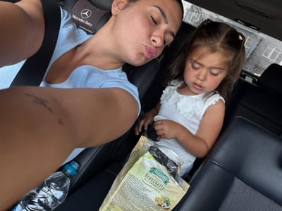 In a car, a woman with closed eyes and puckered lips is taking a selfie while a young girl looks down at a bag of Marretti Bruschette Chips. The woman is wearing a light blue top and a black seatbelt. A Mercedes-Benz headrest is visible behind her. The girl is wearing a white top with floral embellishments. A water bottle is on the seat between them. The background shows a blurry, rainy cityscape, likely Struma, Bulgaria, with buildings and trees visible through the car's windows.