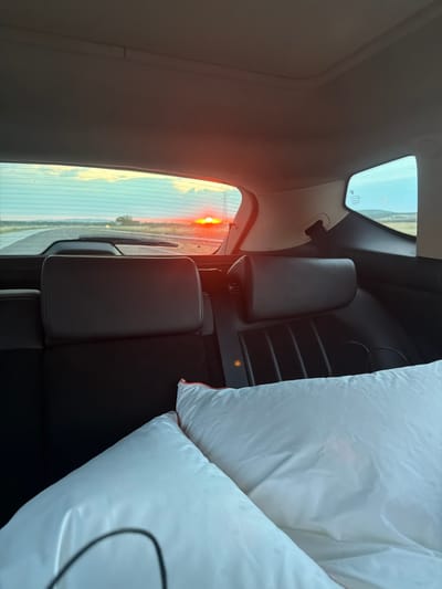 This is a rear interior view of a car, looking out the back windshield at sunset. Two dark-colored headrests of the car's back seats are prominently visible in the foreground, with two white pillows laid out on the seats in front of them. The pillows are smooth and appear to be for sleeping. Outside the car, a vibrant orange sunset is visible on the horizon, casting a warm glow on the sky and the road ahead. The road appears to be a highway, with a slight curve to the left. There are no people visible in the image. The overall scene suggests a road trip or travel during sunset. The location is stated as Georgi-Dobrevo, Bulgaria.