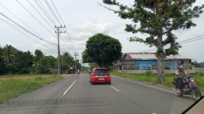 This is a medium-angle, exterior, daytime, full shot of a rural road in Keudealuerheung, Indonesia. The road is asphalt with a yellow lane marker and a white line on the left. On the left side of the road is a field with tall grass, followed by trees and utility poles with wires. On the right side of the road is a two-story building with a corrugated metal roof, blue walls, and a wooden base. A large tree is in front of the building. A red car is driving down the road in the center of the frame, and a motorcycle with a rider is passing the car on the right. The sky is overcast.