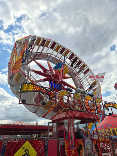 The image captures a vibrant scene at a fairground in Babergh, United Kingdom. The central subject is an amusement ride, a rotating structure with passenger compartments positioned along its circumference. People are visible in the ride, appearing to be enjoying the experience. The ride is adorned with numerous illuminated lights, and banners and flags are hanging from it. To the right side of the frame, there is a partial view of a "Moon Walk" inflatable structure, with additional fairground attractions situated behind it. The environment includes a sky filled with clouds. A "KEEP OUT" sign is visible on one of the structures. The photograph is taken at an upward angle, showcasing the height of the amusement ride against the backdrop of the sky.