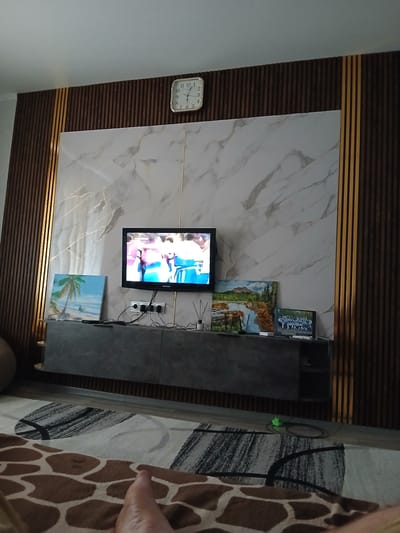 A television is mounted on a wall above a media console in a room. The wall behind the television and console is decorated with a marble-patterned panel. The console itself appears to be made of a grey material. To the left of the television, a framed picture of a landscape with mountains and a river is visible. Above that, another framed picture appears to depict a group of people. A square clock with a white face and black numbers is mounted on the wall to the left of the television. The room's decor includes dark wooden slatted panels on the wall above the marble panel. To the right of the console, a rug with a giraffe-like pattern is visible. The lighting suggests it is daytime. The setting appears to be a modern living room. There is no visible text in the image.