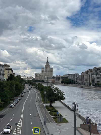 The image shows a cityscape with a prominent building in the distance, likely one of the Seven Sisters skyscrapers in Moscow, Russia. A wide road runs through the scene, lined with trees and parked cars. A river flows parallel to the road on the right side of the frame, with buildings lining its bank. The sky is cloudy with patches of blue. Streetlights, road markings, and signs are visible along the road. Pedestrian infrastructure such as walkways and crosswalks are also present. There is a crosswalk symbol. There are no people visible in the image. Sertyakino, Russia is not featured in the image.