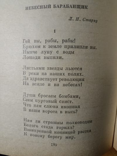 The image is a close-up, high-angle shot of an open book, displaying a poem in Russian. The text is printed in a serif font. The title of the poem, "НЕБЕСНЫЙ БАРАБАНЩИК" (Heavenly Drummer), is centered at the top, followed by the author's name, "Л. Н. Старку" (L. N. Starku), also centered. Below this, the number "1" is displayed, indicating it is the first part or stanza. The poem itself consists of several stanzas of verse. At the bottom of the page, the number "180" is present, likely a page number. The book's pages appear to be aging, with a slightly yellowed and textured appearance. The lighting suggests an indoor setting. The location of Dagestanskiye Ogni, Russia is not depicted or mentioned in the image.
